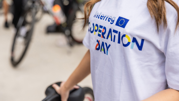 Person wearing a white T-shirt with "Interreg OPERATION DAY" in colorful letters. They are holding a bicycle seat, and blurred bicycles are visible in the background.