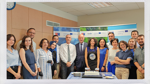 A group of fifteen people, both men and women, stand around a table with a cake in an office setting. Most are smiling and dressed in business casual attire. Banners and a clock are visible in the background.